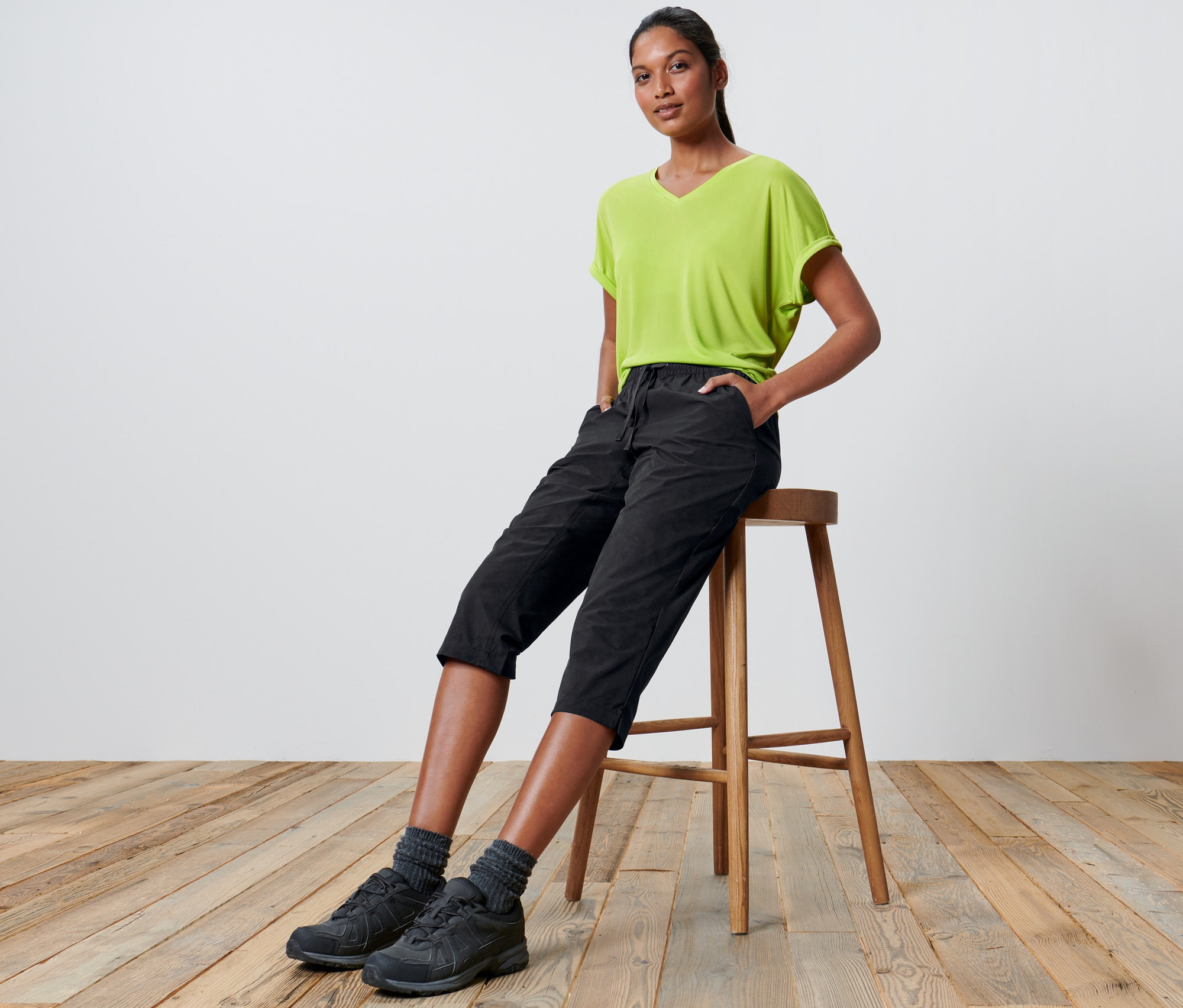 Femme assise sur un tabouret portant un pantacourt technique en softshell et un t-shirt vert.
