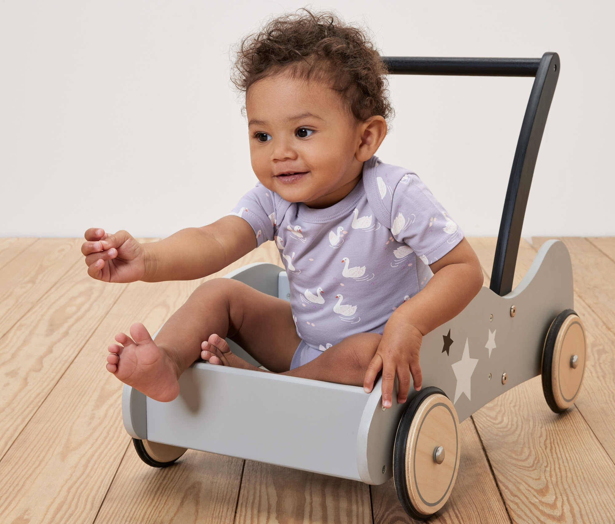 Un bambin aux cheveux bouclés est assis dans un chariot de marche en bois gris avec un motif d'étoiles et porte un lot de 5 bodys évolutifs pour bébé princesse.
