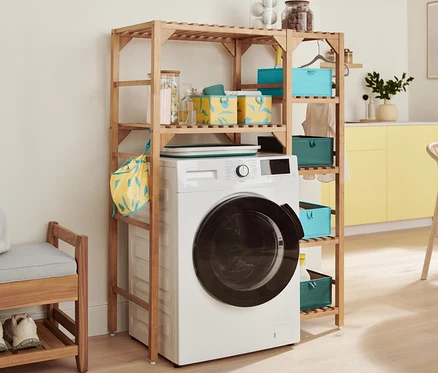 Une machine à laver blanche est placée sous une étagère en bois. Sur l'étagère se trouvent des boîtes de rangement pour tiroir, un panier à linge pliable et une étagère pour machine à laver. À gauche se trouve un banc avec espace de rangement.