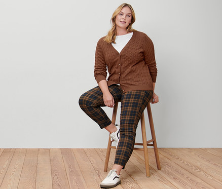 Une femme est assise sur un tabouret et porte un cardigan en maille fine à motif torsadé, un t-shirt manches 3/4 blanc, un pantalon stretch à carreaux et des baskets en cuir.