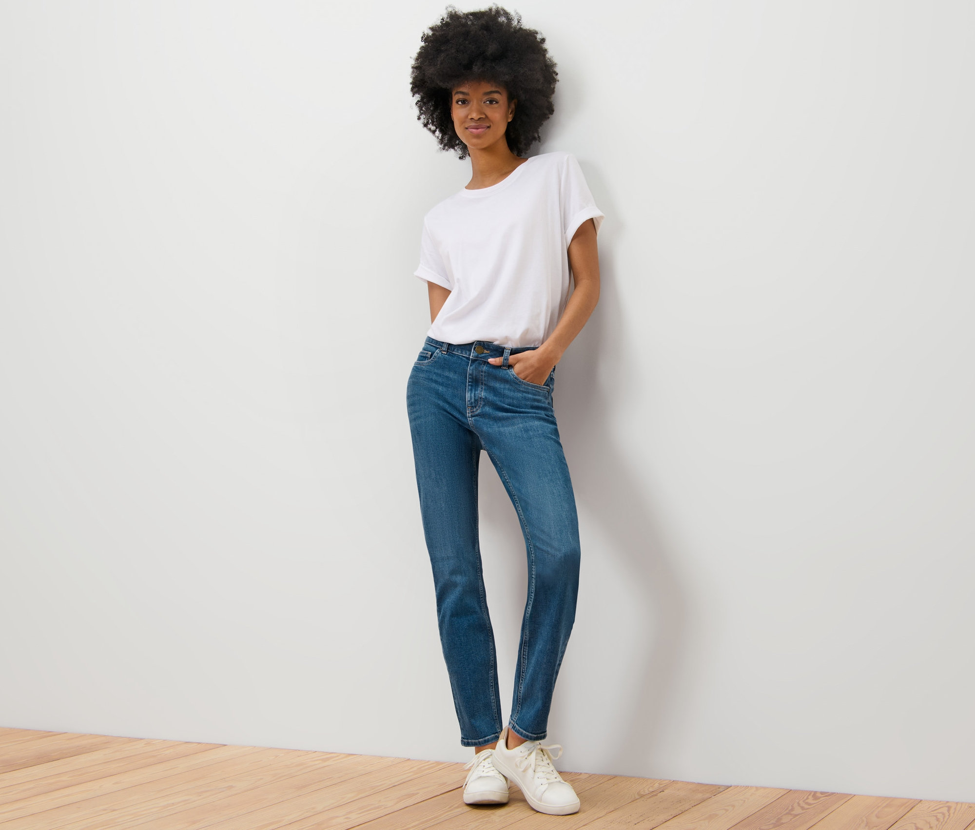 Une femme pose contre un mur, elle porte un jean slim – Coupe « Emma » bleu moyen, un t-shirt blanc et des baskets.