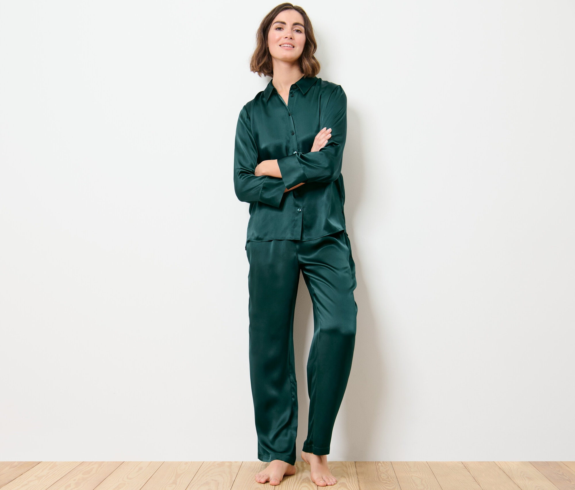 Une femme se tient debout, les bras croisés, portant un pyjama en soie vert foncé. Son haut de pyjama en pure soie est boutonné.