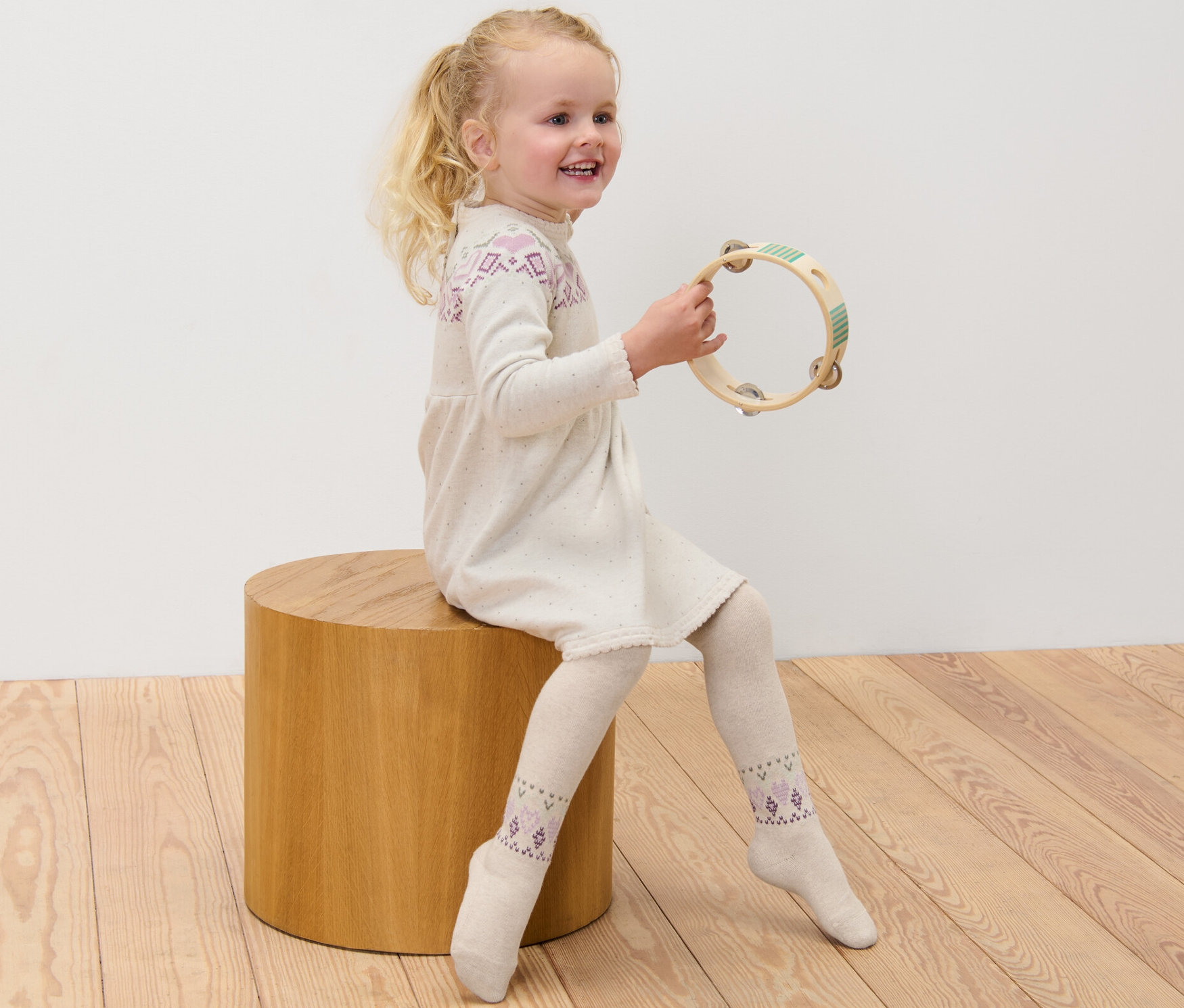 Une fillette est assise sur un tabouret en bois et joue du tambourin. Elle porte une robe et 3 collants pour enfant.