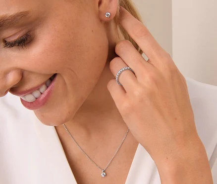 Une femme souriante touche son oreille, portant une bague argent 925 mémoire, un collier en argent 925 solitaire et des clous d’oreilles en argent 925 solitaire.
