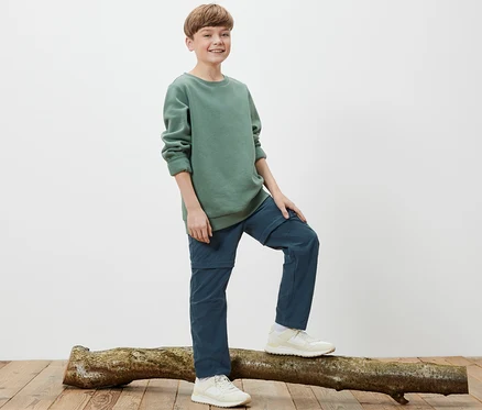 Un garçon se tient sur un tronc d'arbre et porte un pantalon fonctionnel pour enfant.