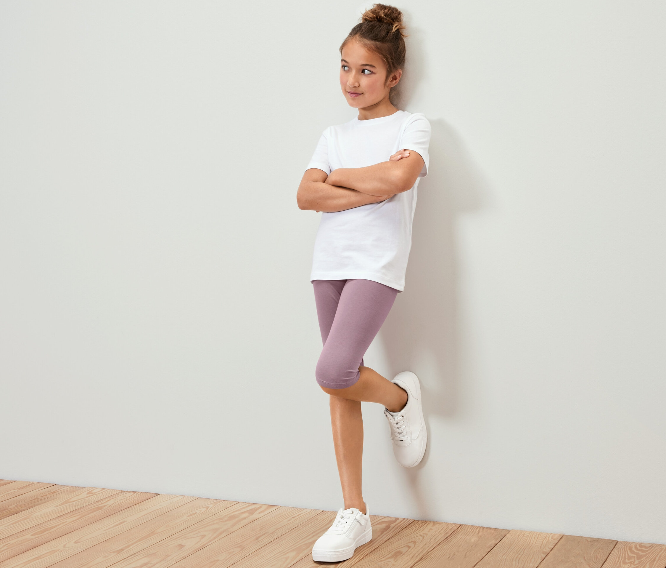 Une fille portant un t-shirt blanc et un legging 3/4 violet (3 leggings 3/4 pour enfant) est appuyée contre le mur.
