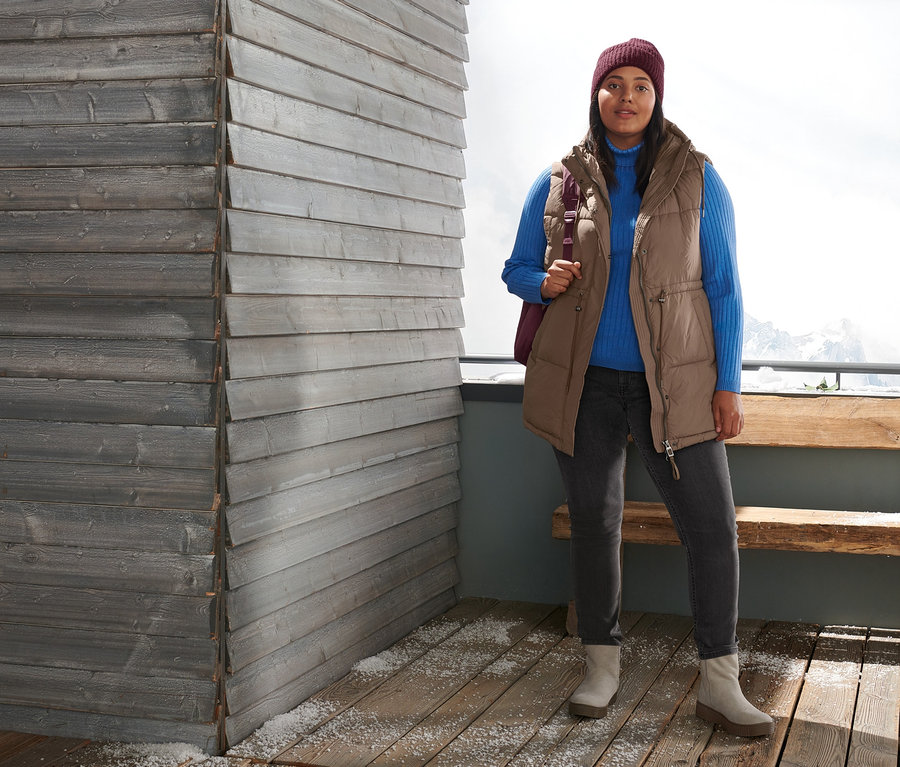 Une femme se tient à l'extérieur et porte un gilet matelassé, un pull à col roulé côtelé, un jean slim, des bottines en cuir velours et un bonnet en maille rouge bordeaux. Elle porte un sac à dos matelassé pour l'extérieur.