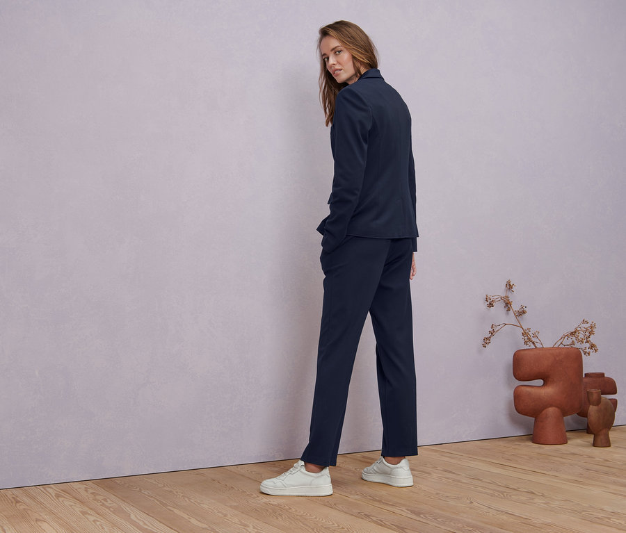 Une femme regarde par-dessus son épaule et porte un blazer en jersey bleu foncé, un pantalon en jersey avec nervure bleu foncé et un t-shirt à demi-manches blanc.