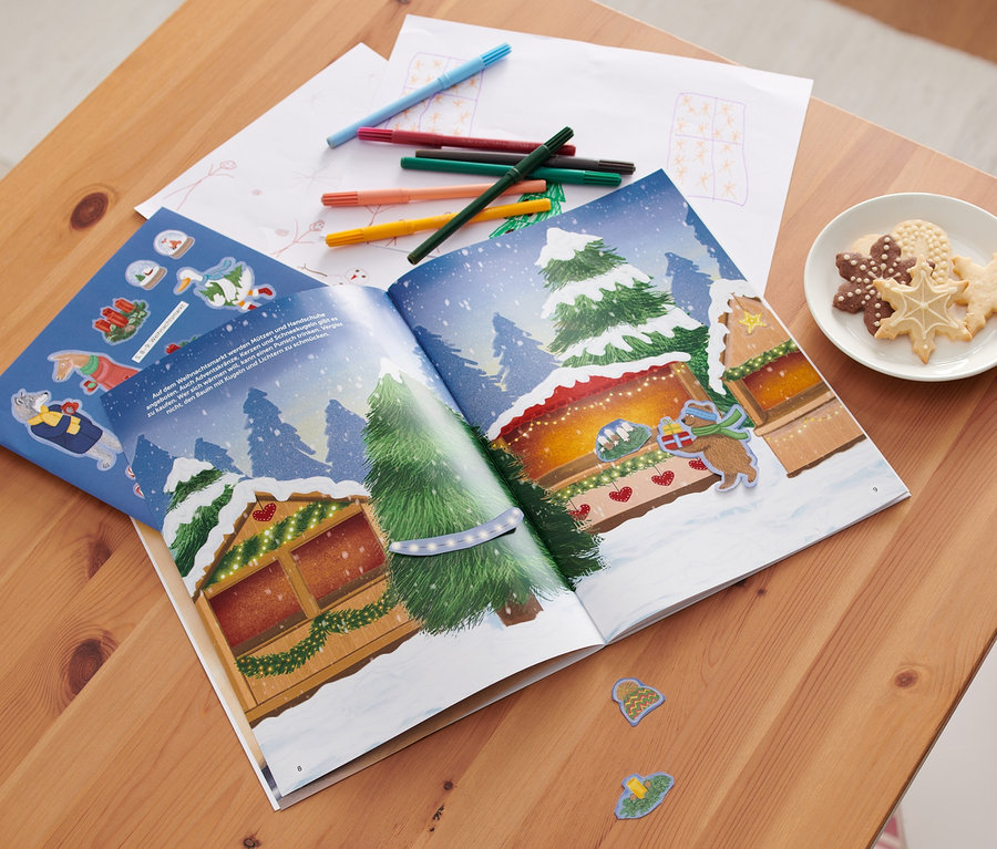 Sur une table en bois, un livre d'autocollants ouvert : « Bezaubernde Weihnachtszeit » avec un motif de Noël. À côté, des biscuits, des crayons et un renard en peluche.