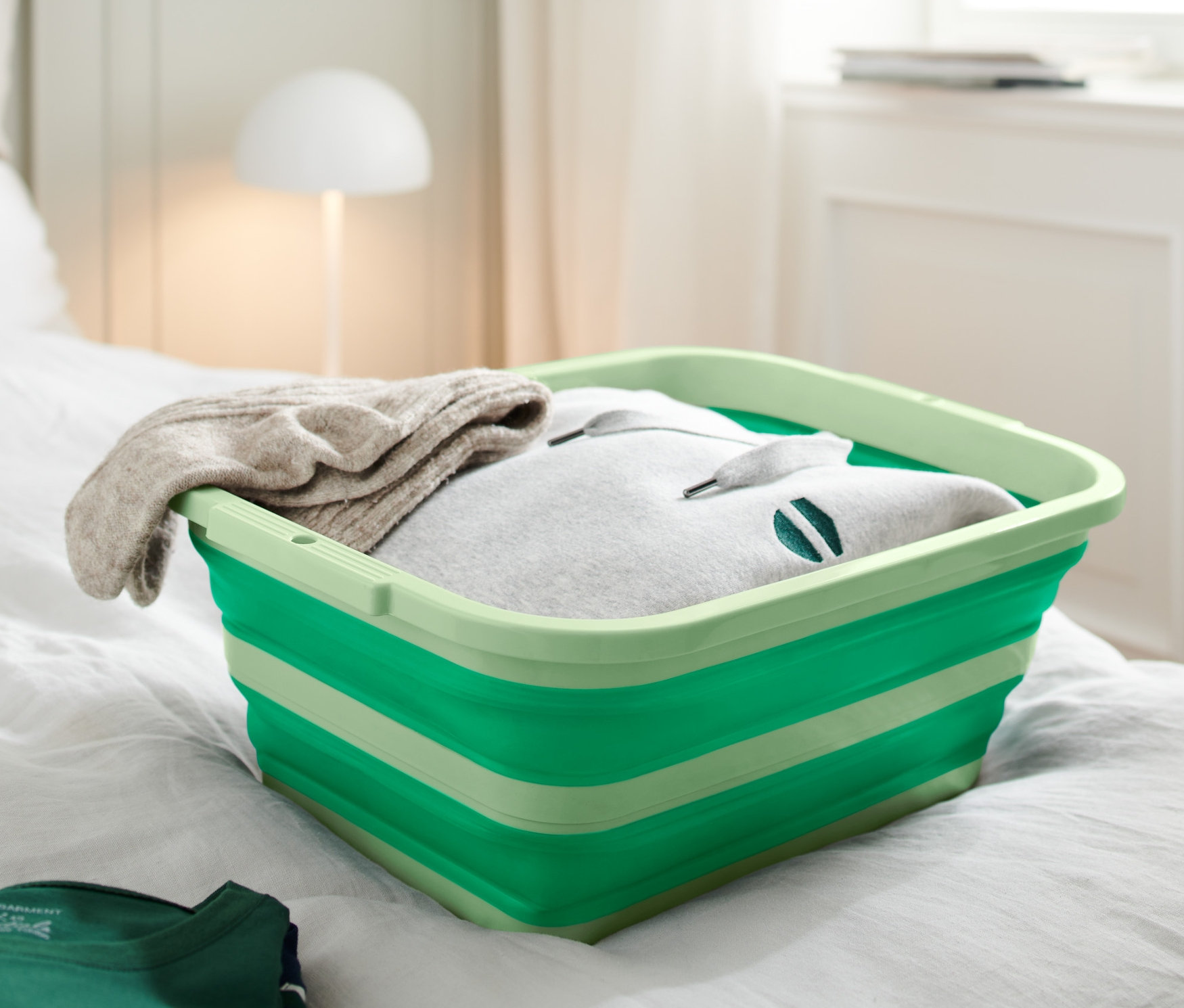 Un panier à linge et à vaisselle pliable vert est posé sur un lit. Le panier est rempli de vêtements.