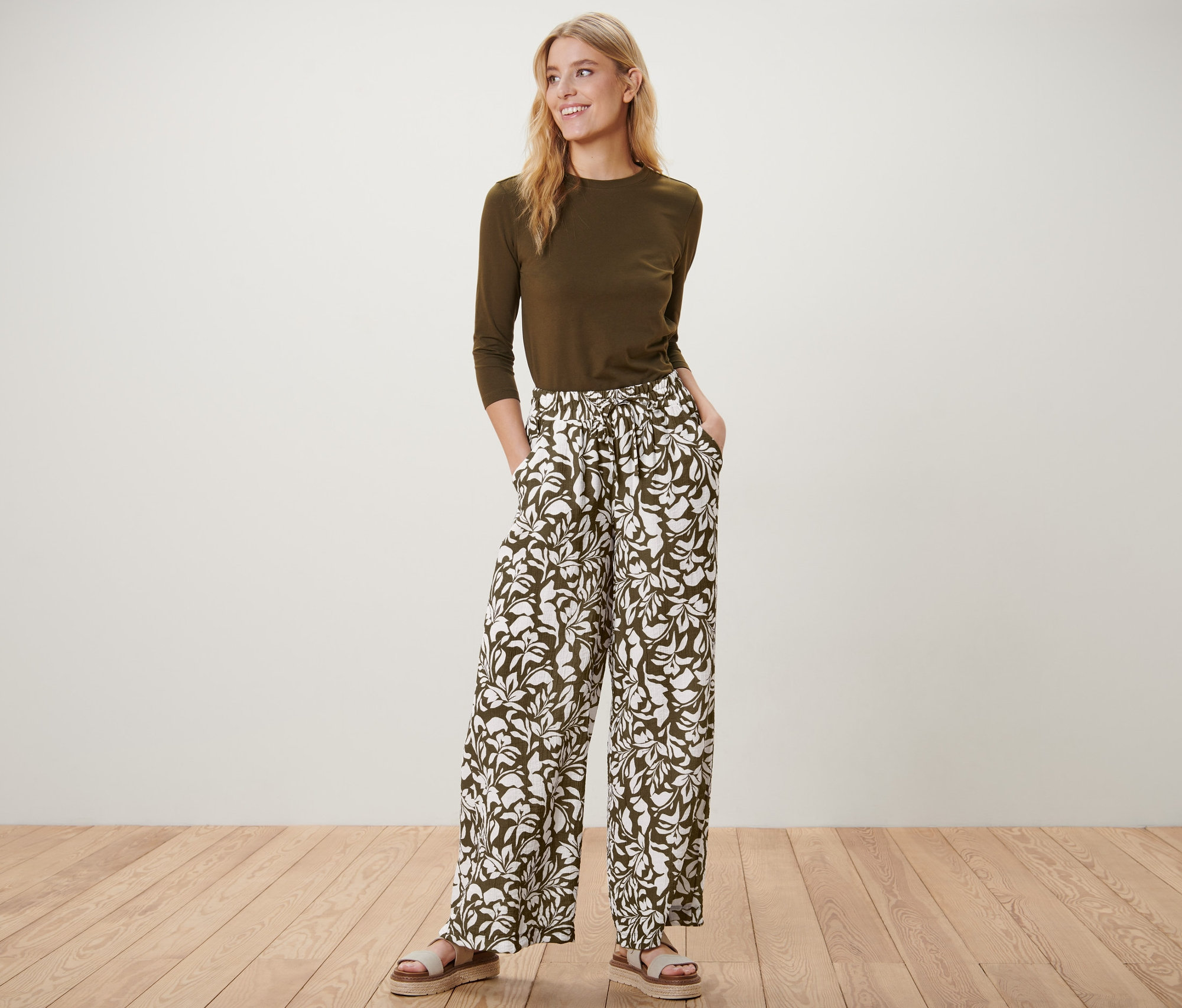 Une femme pose debout portant un pantalon imprimé à taille élastique, un haut olive et des sandales.