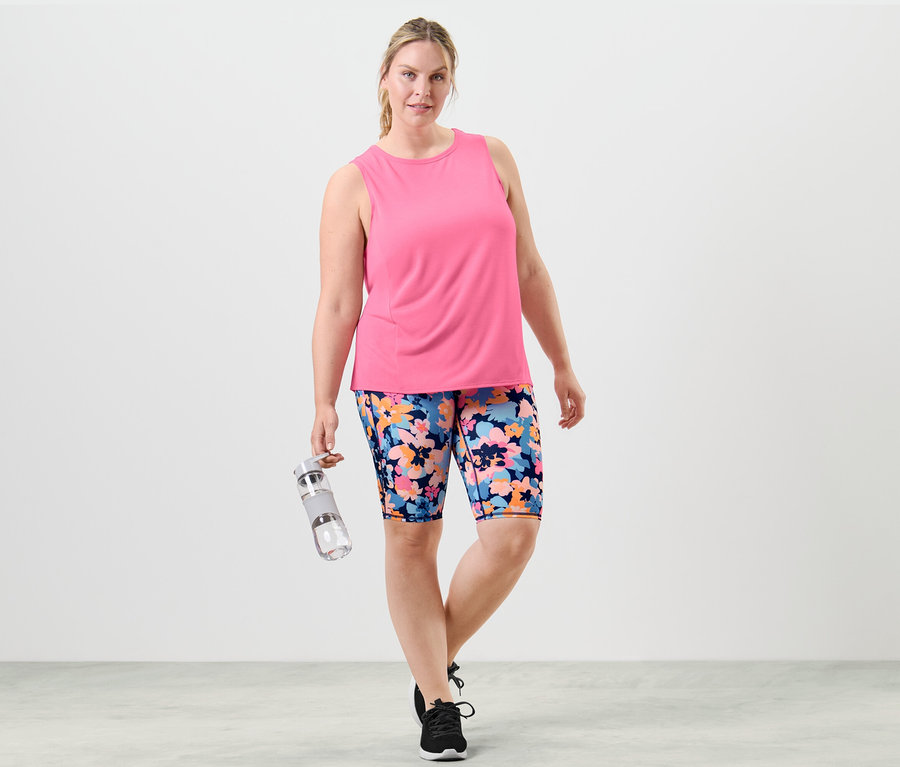 Une femme pose en tenue de sport, portant un top pêche et un short à fleurs, tenant une bouteille d'eau.