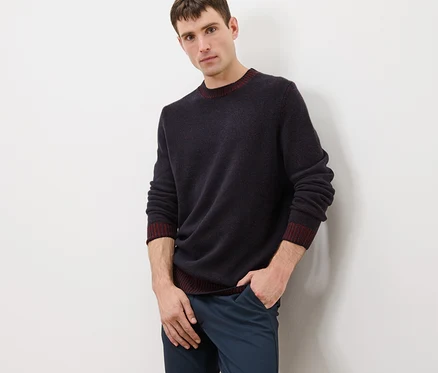Un homme pose en pull en laine bleu marine avec des finitions bordeaux et un pantalon bleu.
