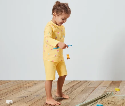 Une fille joue sur une couverture avec du sable, portant une combinaison jaune pour enfants avec un indice de protection UV 80.