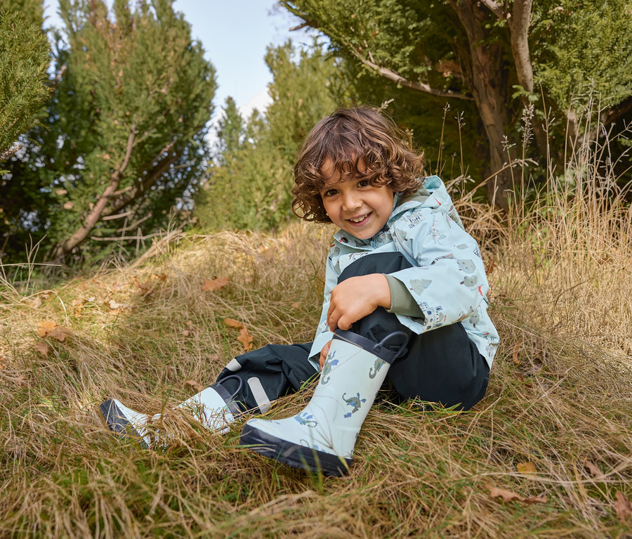Un garçon est assis dans l'herbe et porte des bottes de pluie pour enfant, dragon, une veste de pluie pour enfant, chevalier et un pantalon de pluie pour enfant.
