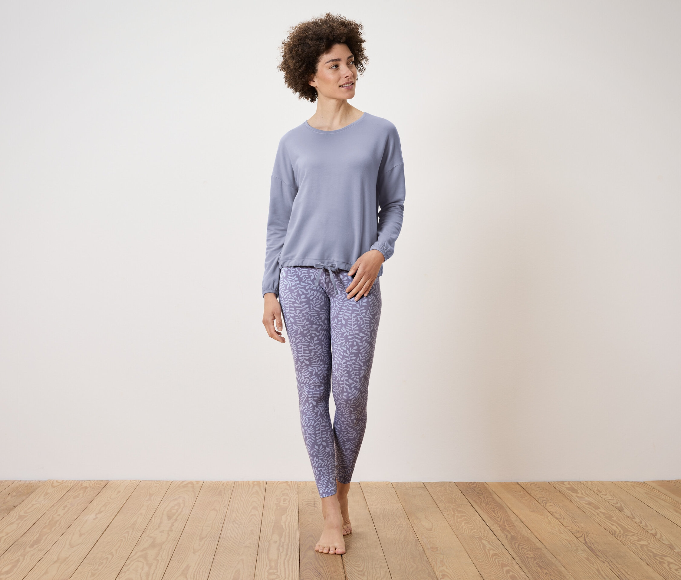 Femme en pyjama bleu clair avec motif de feuilles debout sur un plancher en bois.