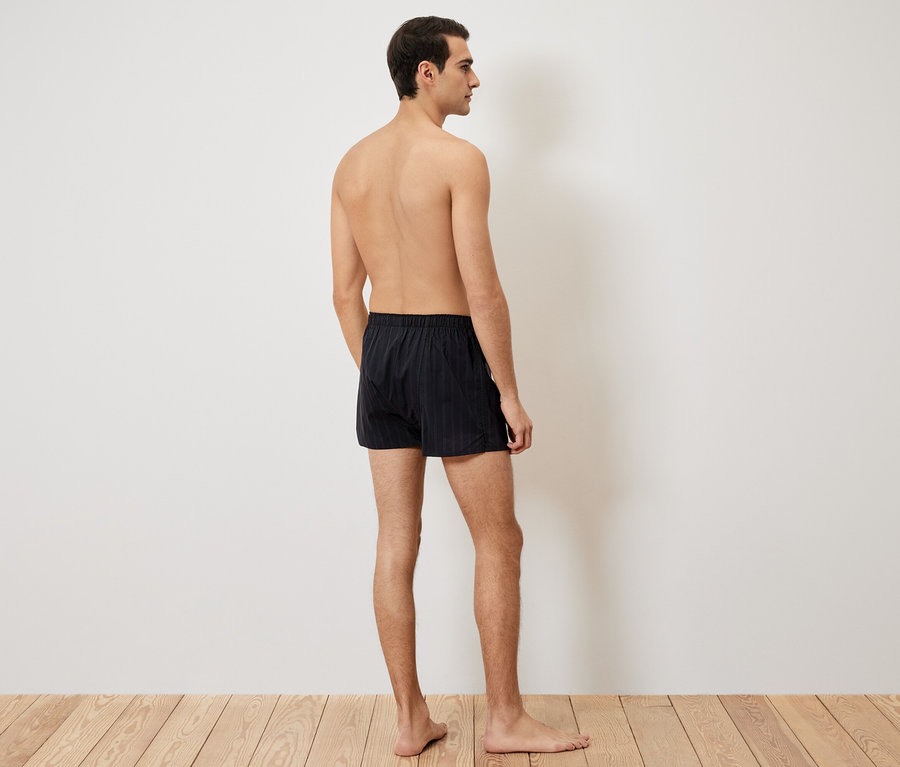 Homme vu de dos, portant un caleçon boxer noir sur un plancher en bois.