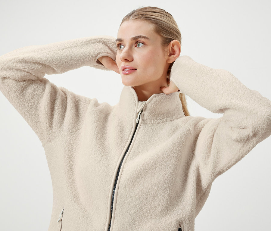 Une femme blonde aux cheveux tirés en queue de cheval pose en veste polaire crème zippée.