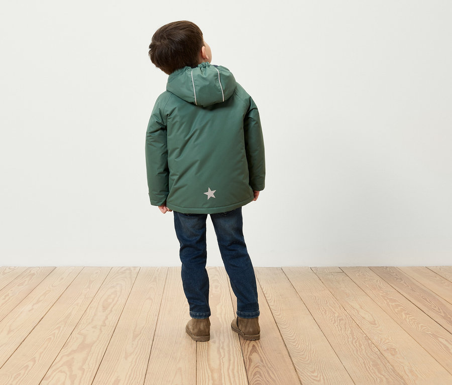 Un garçon vu de dos porte un manteau d'hiver vert à capuche, un jean bleu et des chaussures marron, debout sur un plancher en bois.