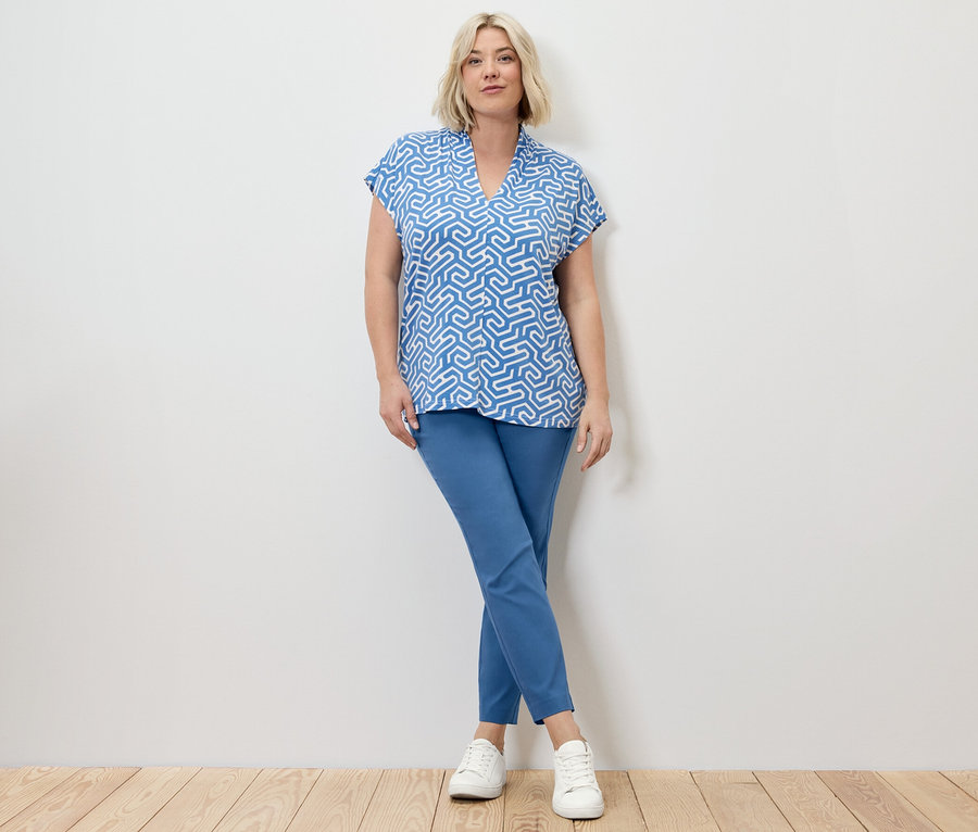 Une femme blonde pose en pantalon stretch et chemisier à col en V, tous deux bleus.