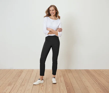 Une femme pose, les bras croisés, en pantalon marron et chemise blanche.