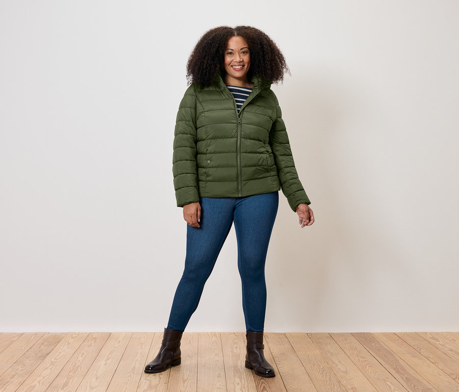 Une femme debout porte une doudoune vert olive, un jean et des bottes marron.