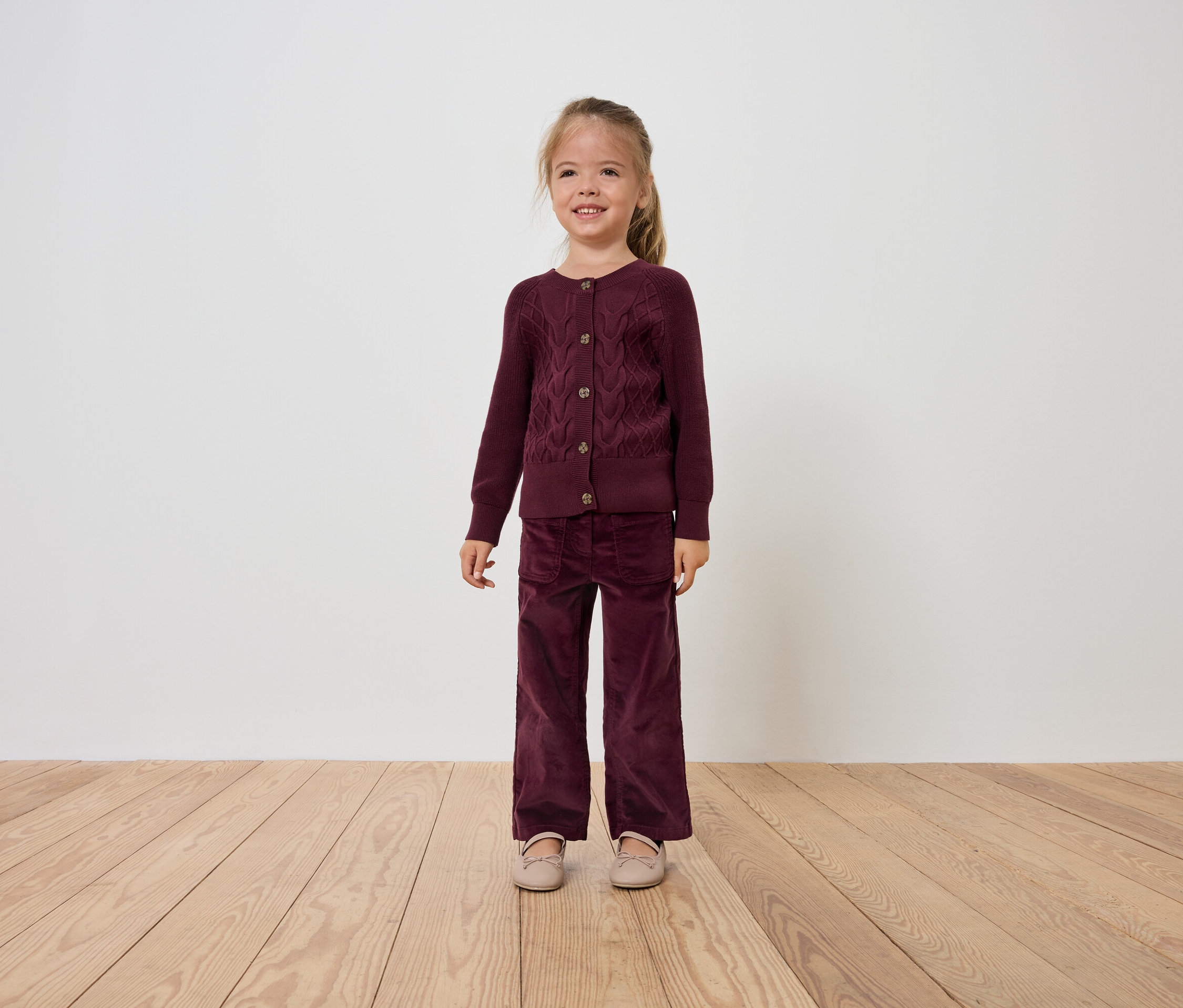 Une fillette souriante vêtue d'un cardigan et d'un pantalon en velours bordeaux se tient sur un plancher en bois.