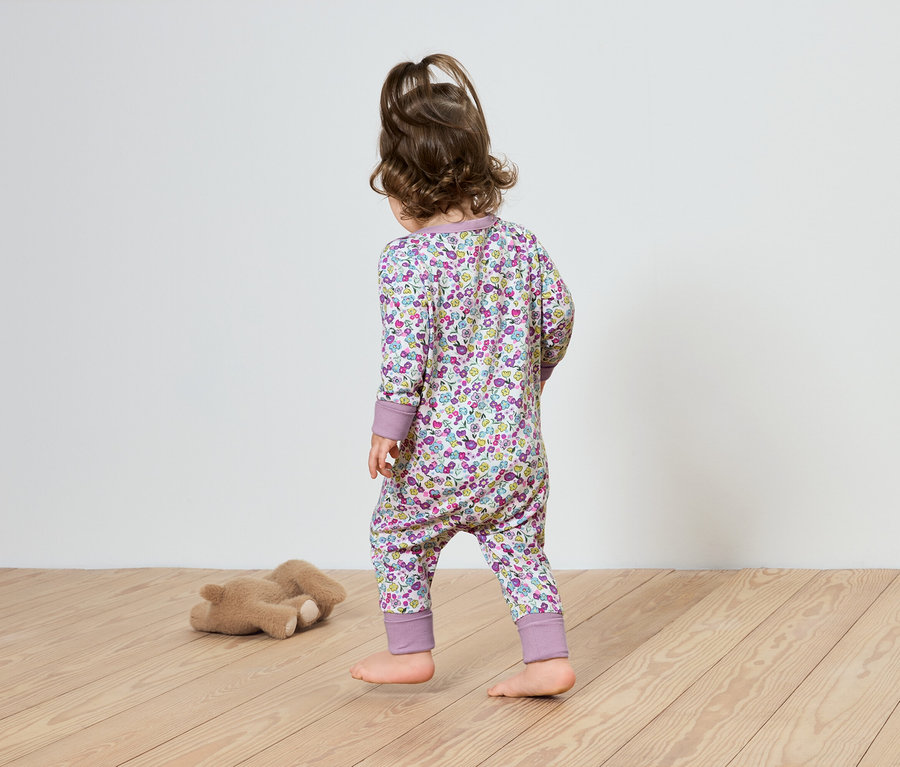 Un enfant en combinaison fleurie marche sur un plancher en bois en direction d'un ours en peluche.