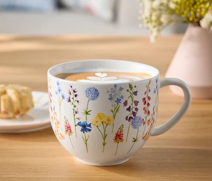 Tasse à fleurs avec café et cœur de latte art sur une table en bois à côté d'une assiette de pâtisseries et d'un vase de fleurs.