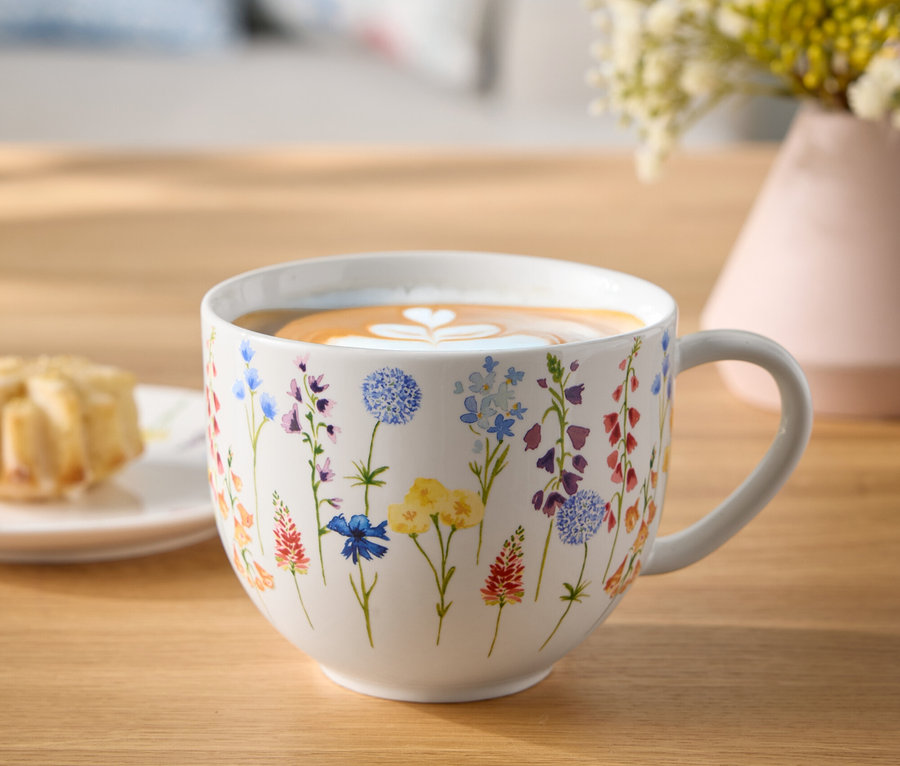Tasse à fleurs avec café et cœur de latte art sur une table en bois à côté d'une assiette de pâtisseries et d'un vase de fleurs.