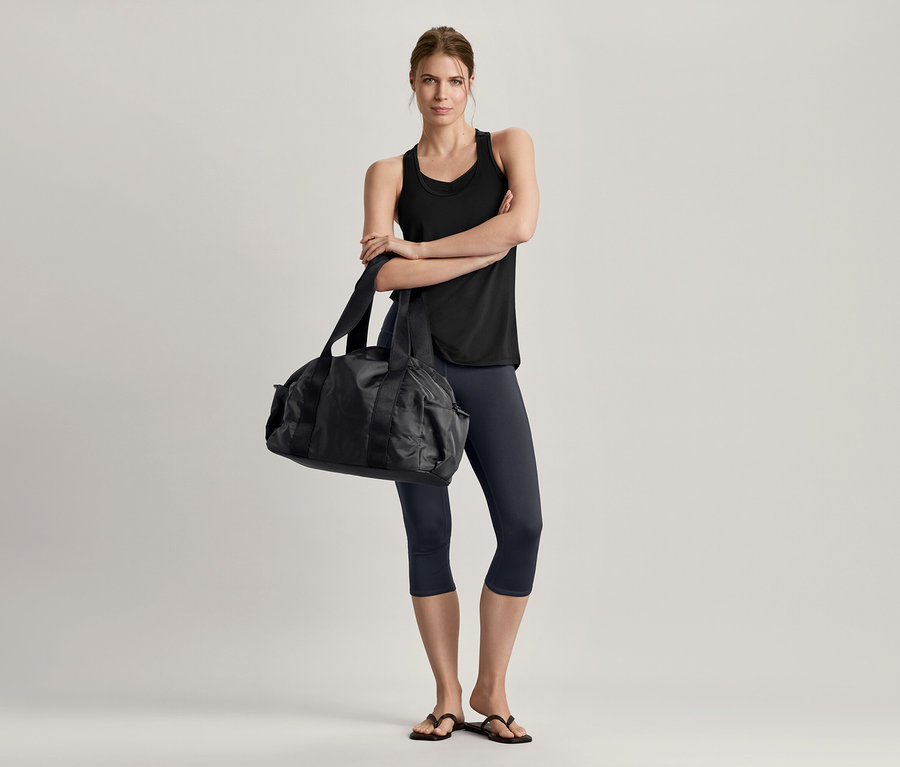 Femme les bras croisés portant un bustier de sport sans coutures noir, un legging de sport 3/4 et tenant un sac de sport noir.
