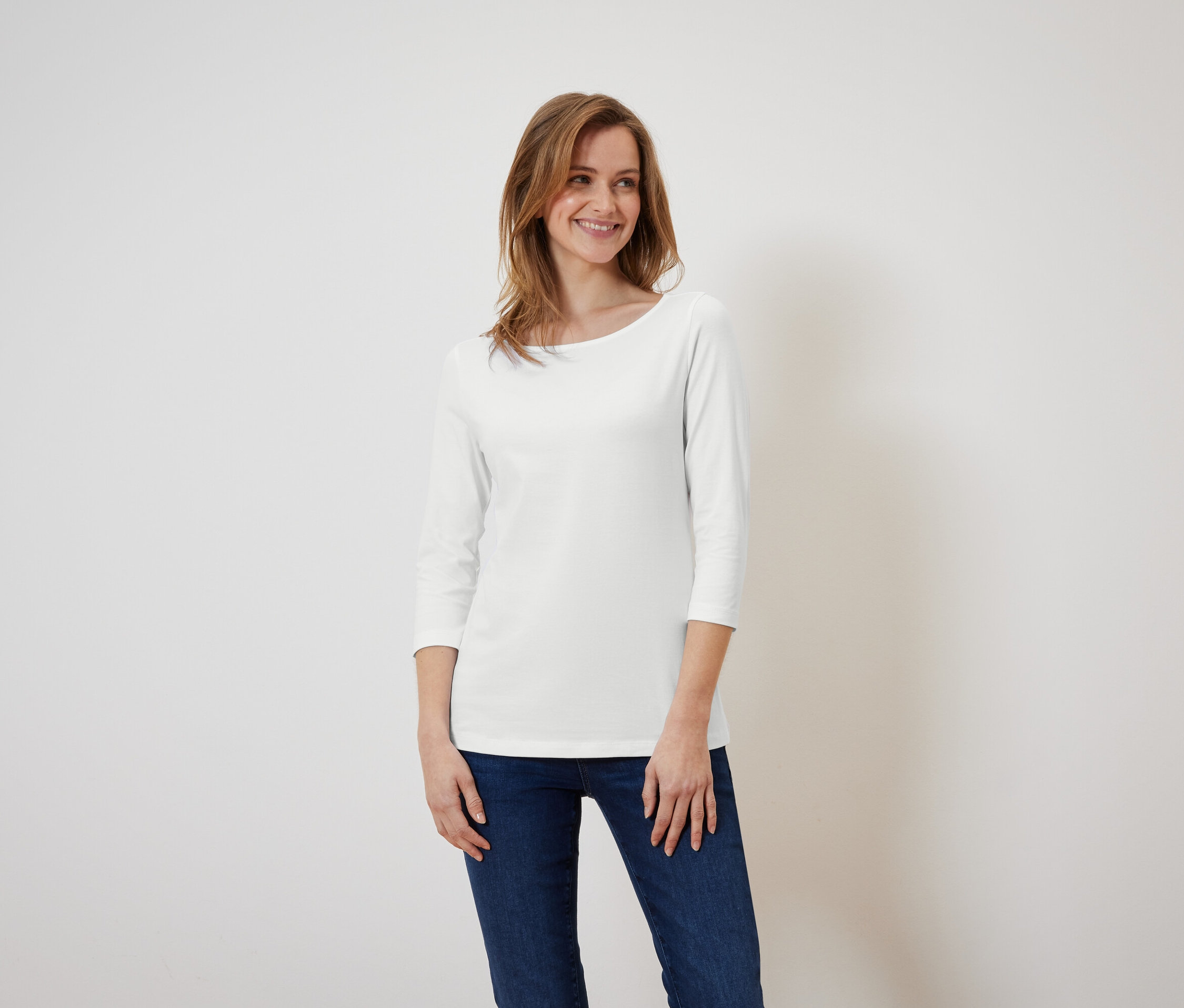 Une femme souriante pose devant un fond blanc, portant un t-shirt blanc à manches trois-quarts et un jean bleu.