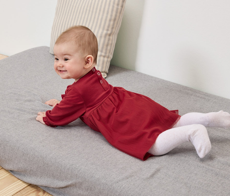 Bébé en robe rouge et collants blancs allongé sur un tapis gris.