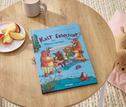 Sur une table en bois reposent le livre "Kalt Erwischt", une assiette de quartiers de pommes, une tasse et un chien en peluche sur un coussin rose.