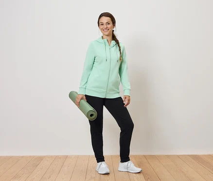 Une femme tient un tapis de yoga vert enroulé, porte un sweat à capuche vert menthe et un pantalon de survêtement noir.