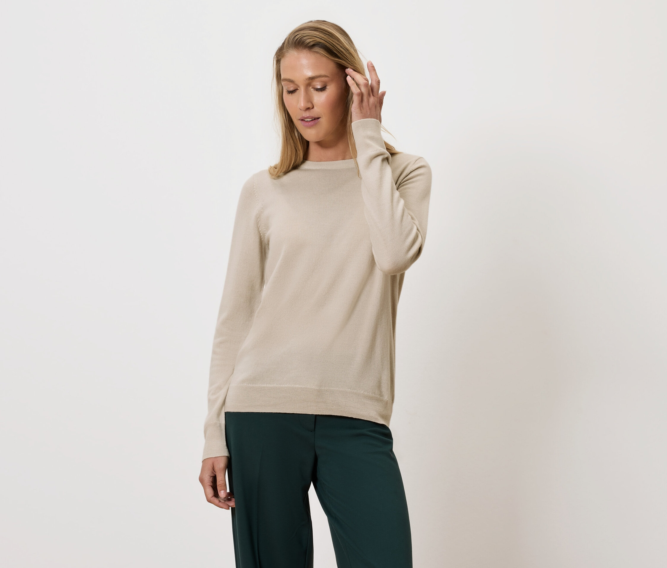 Une femme portant un pull beige et un pantalon vert pose sur un fond clair.