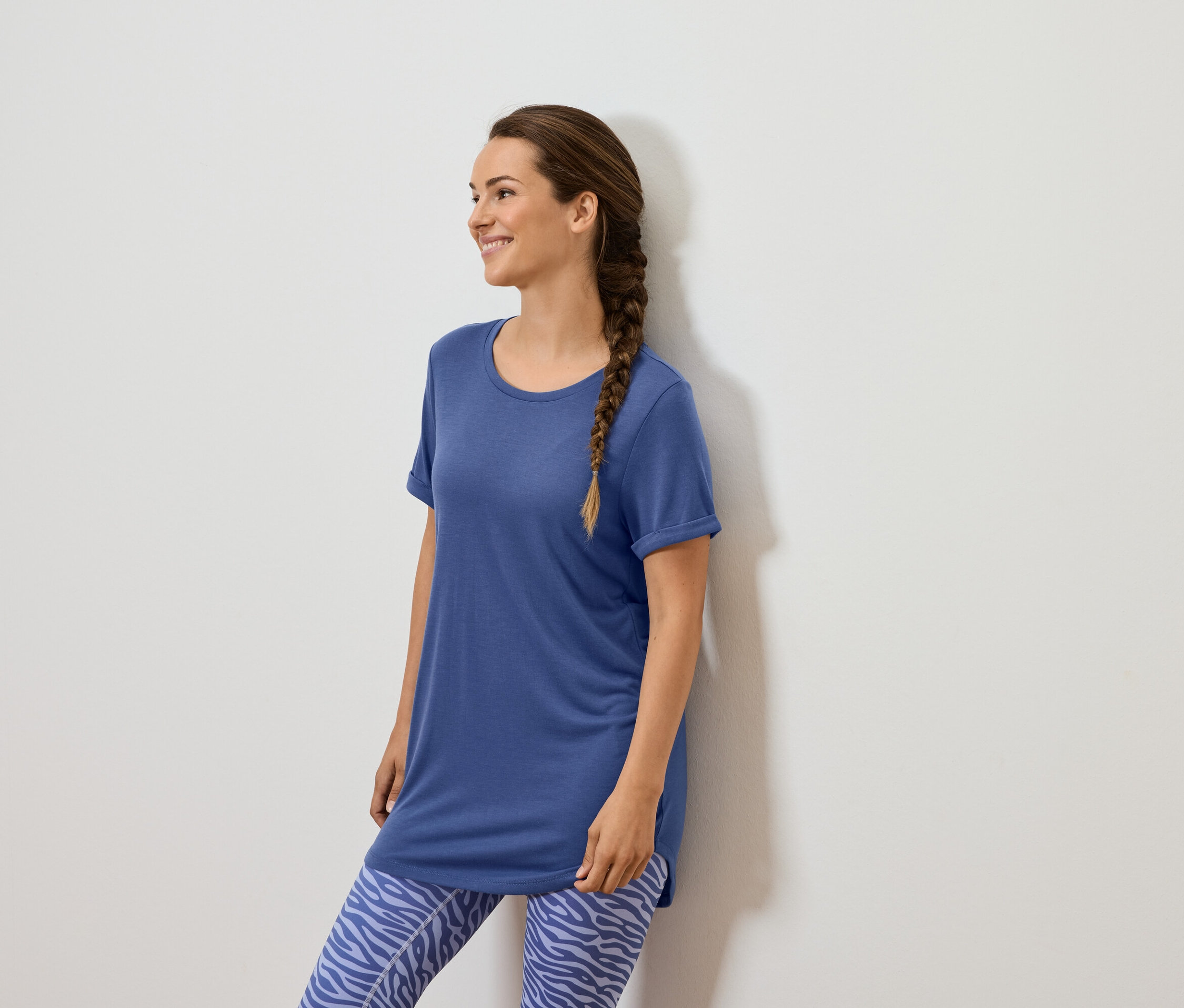 Une femme avec une tresse brune se tient contre un mur blanc, portant un t-shirt bleu et des leggings bleus à motifs animaliers.