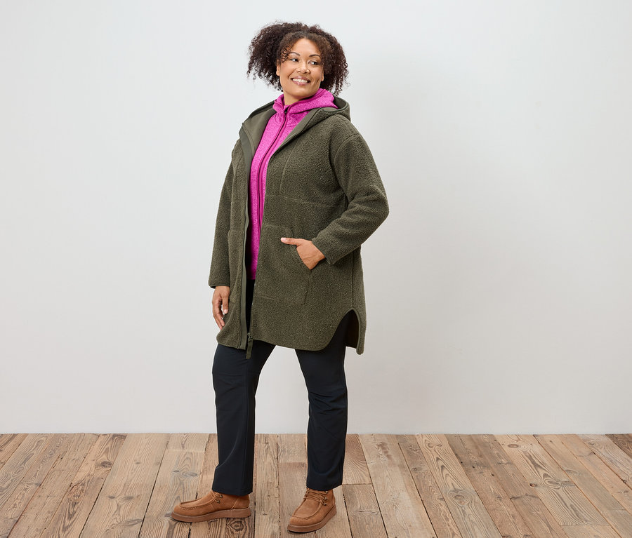 Femme portant un manteau à capuche vert olive, un sweat à capuche rose, un pantalon bleu marine et des chaussures marron, debout sur un plancher en bois.