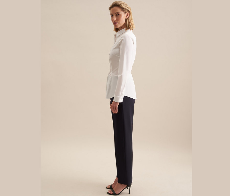 Une femme se tient de côté et porte un chemisier Seidensticker Slimfit blanc et un pantalon bleu marine.