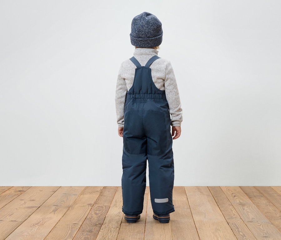 Un garçon, vu de dos, porte une salopette bleue, un pull gris et un bonnet bleu.