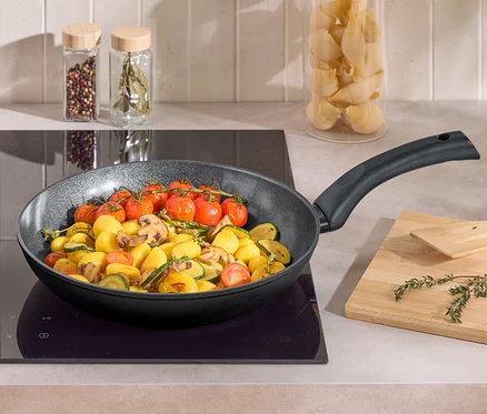 Dans une poêle sur une plaque de cuisson, des pommes de terre, des courgettes, des champignons de Paris et des tomates cerises sont en train de cuire. À côté se trouvent une planche à découper en bois et une spatule.