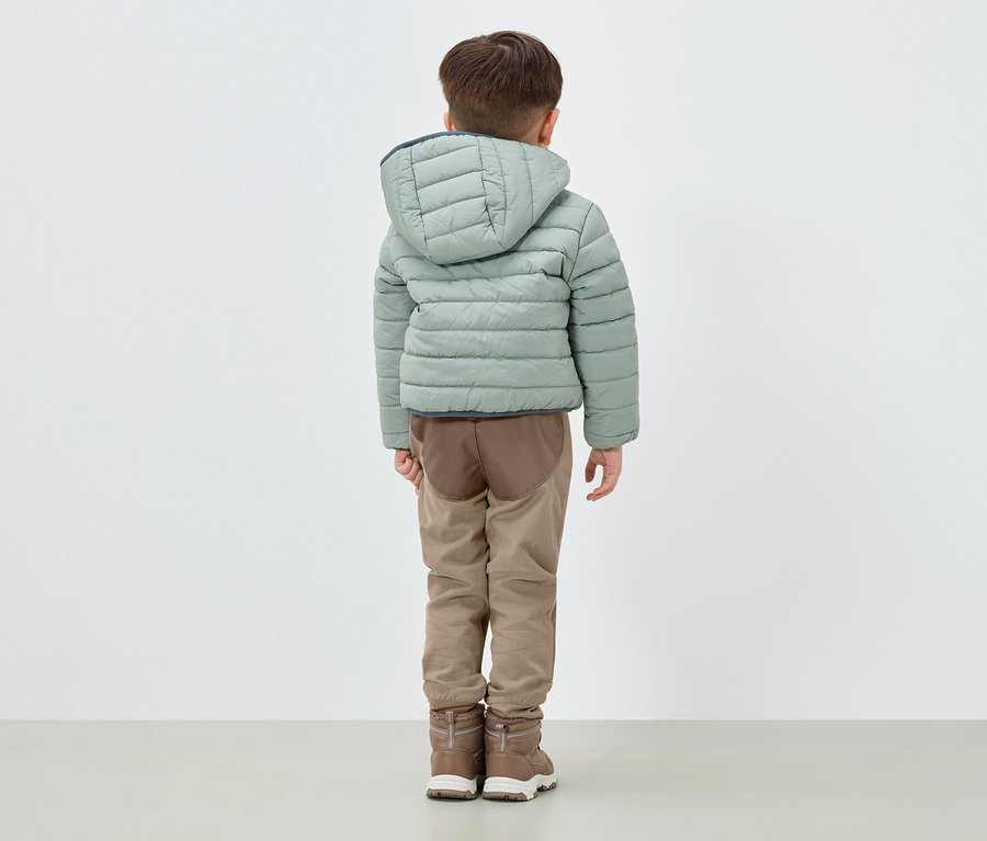 Un garçon se tient dos à l'appareil photo, portant une doudoune vert clair à capuche, un pantalon marron et des bottes marron.