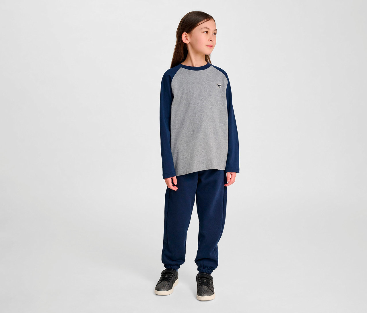 Une fille debout porte un pantalon de survêtement bleu et un t-shirt ample à manches raglan HUMMEL HMLJR BEE gris.