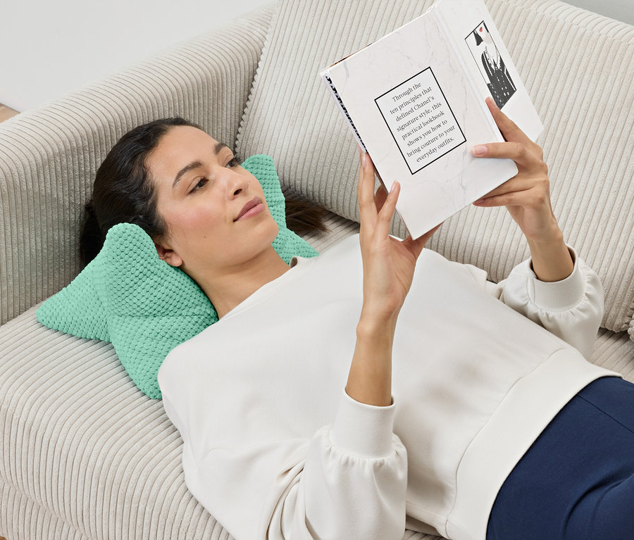 Une femme est allongée sur un canapé et lit un livre, sa tête reposant sur un coussin de lecture.