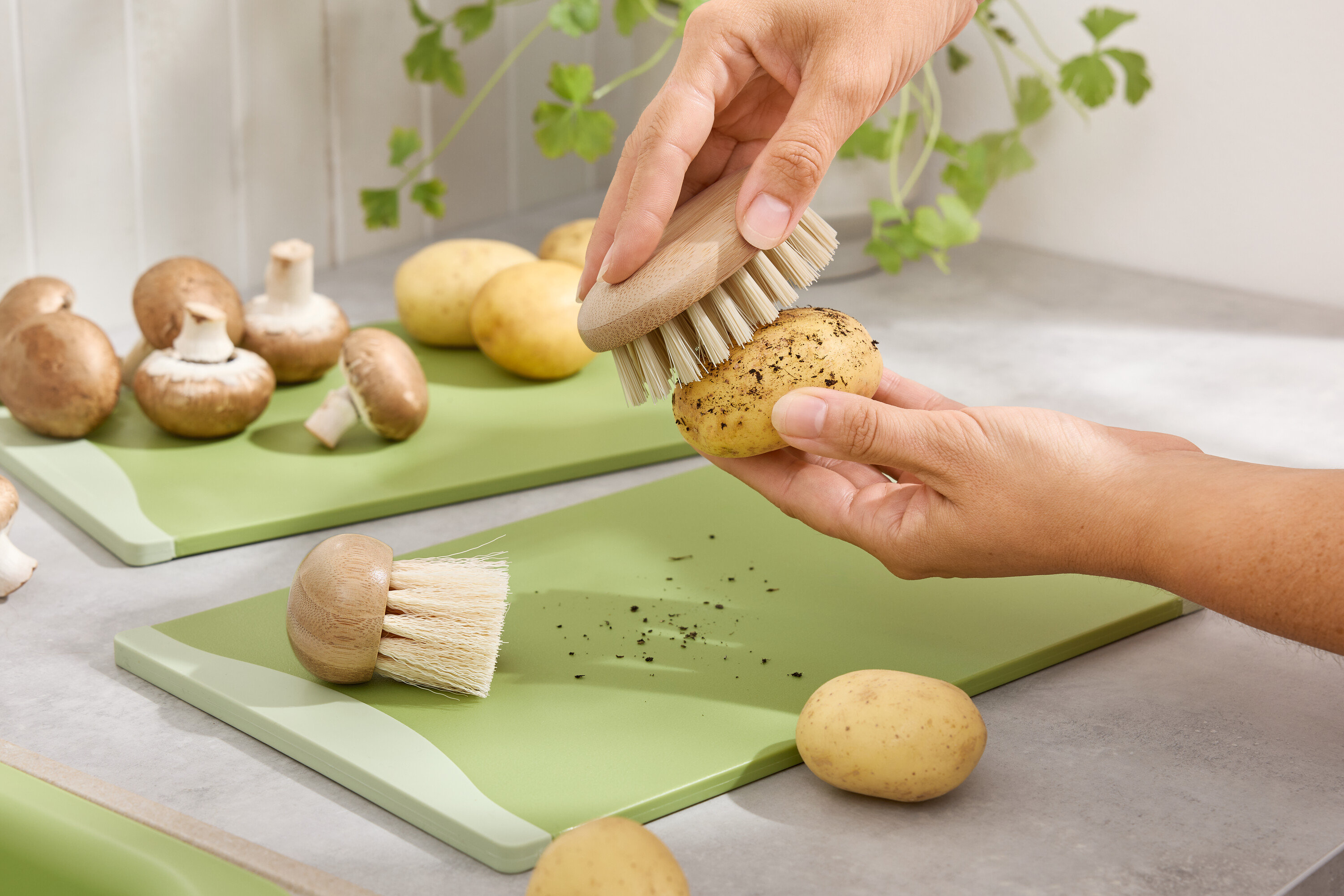 Des mains brossent une pomme de terre sur une planche à découper verte. Des champignons bruns et des pommes de terre sont à l'arrière-plan.