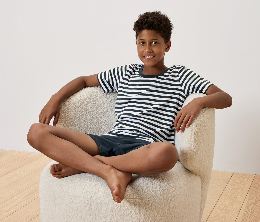 Un jeune homme est assis les jambes croisées dans un fauteuil crème. Il porte un t-shirt rayé et un short bleu marine.