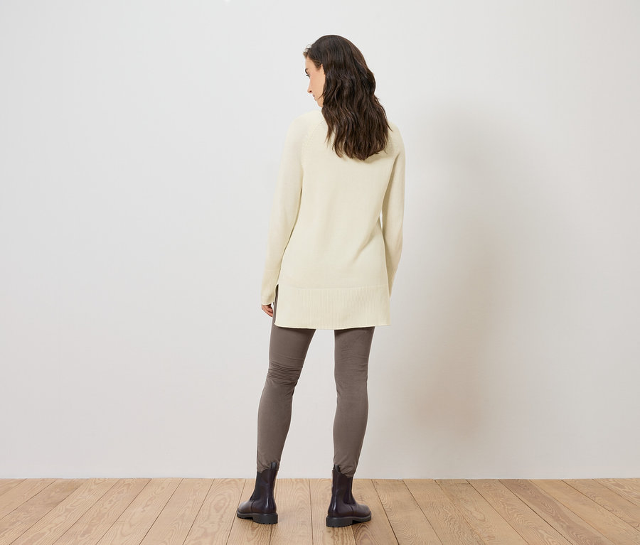 Une femme vue de dos porte un long pull en maille crème, des leggings gris et des bottes noires.
