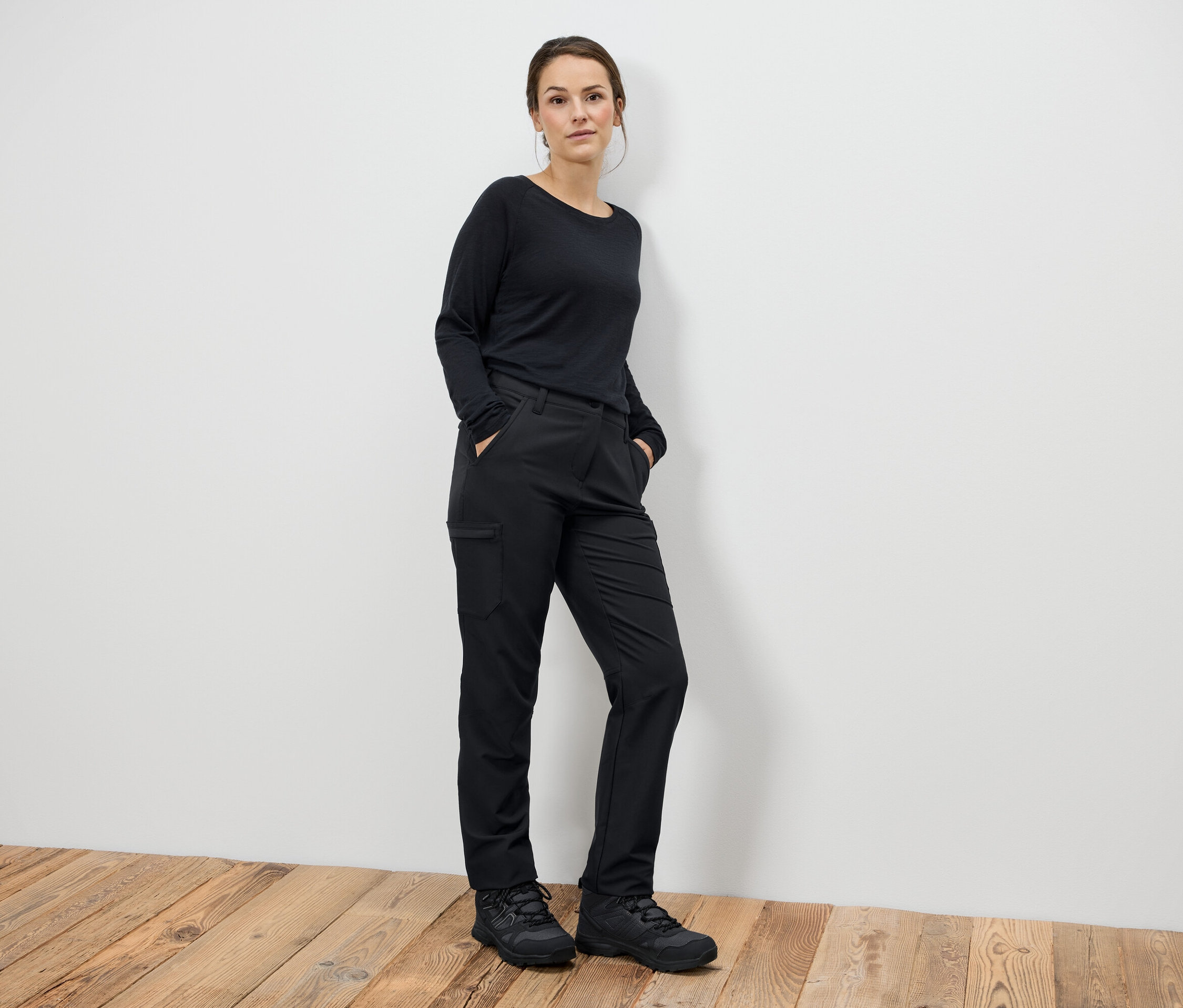 Femme debout, mains dans les poches, portant un pantalon noir et des bottes noires.