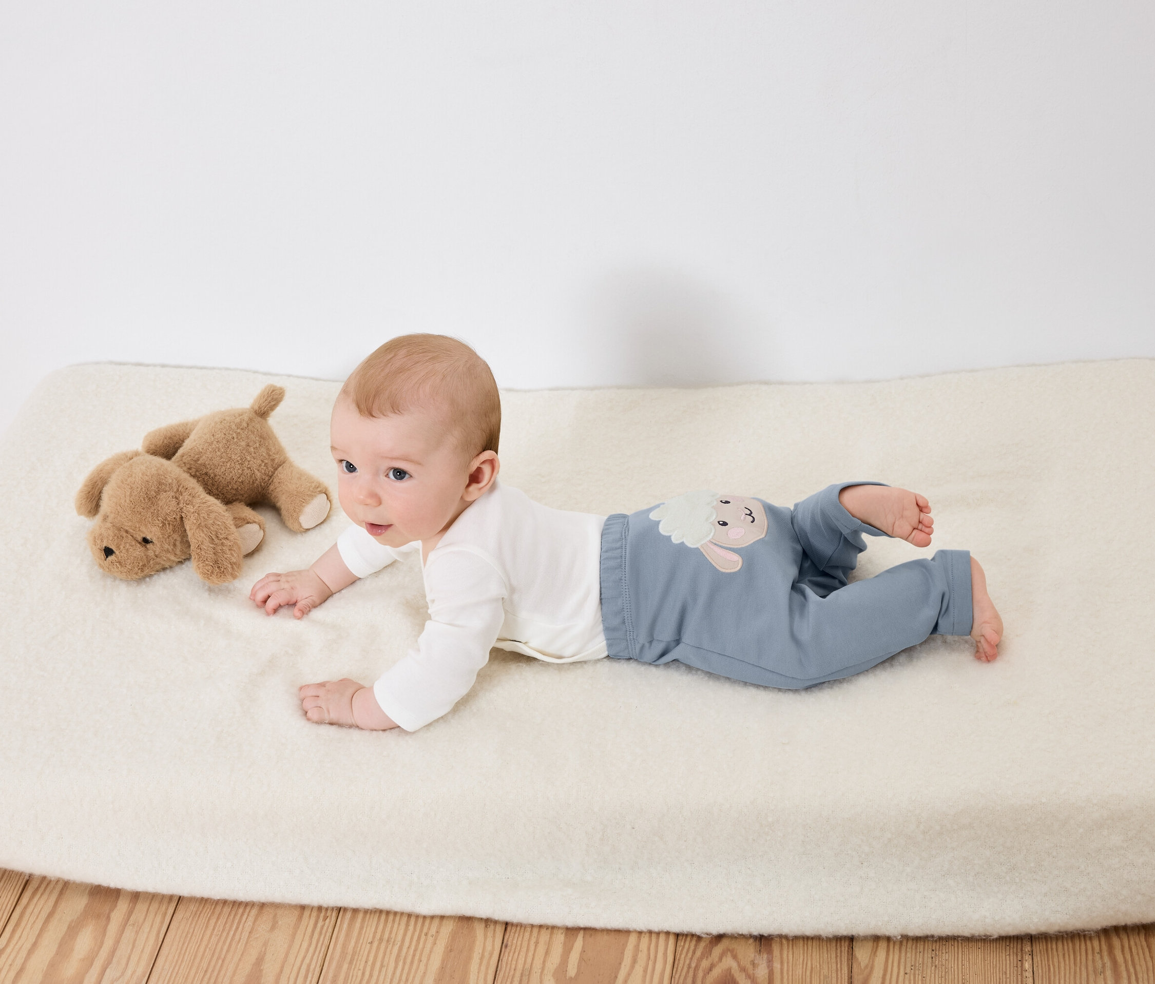 Un bébé est allongé sur le ventre sur une couverture blanche à côté de deux chiens en peluche.