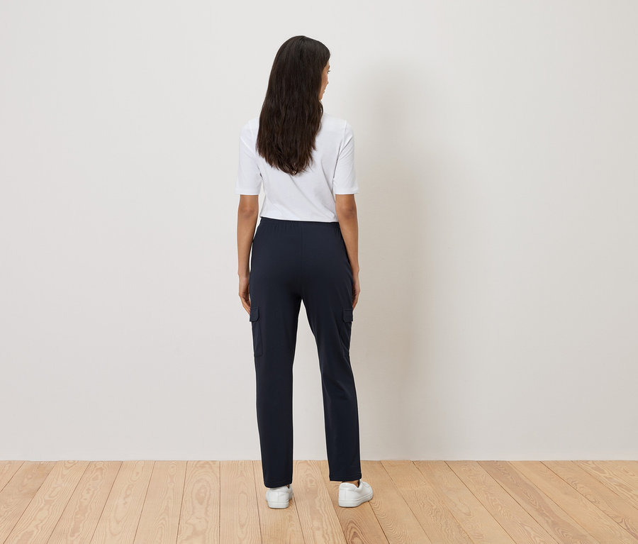 Vue de dos d'une femme aux longs cheveux foncés portant un t-shirt blanc, un pantalon cargo bleu marine et des baskets blanches.